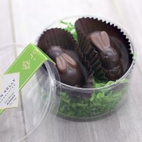 Two bunnies sit in a small, plastic, round, container. There is green crinkle paper beneath them. The lid of the container is learning against the container. The lid features a green label with a John Kelly Chocolates logo.