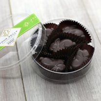 Five dark chocolate bunnies sit in a round, plastic container. The lid of the container is leaned against the container. The lid has a green label on it with a John Kelly Chocolates logo.