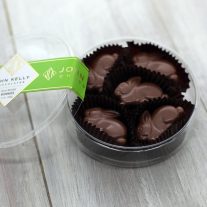 An open container with five milk chocolate bunnies in it. The lid of the container is propped up against the container and has a green label on it with a John Kelly Chocolates logo.