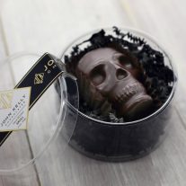 A large dark chocolate skull rests on a bed of black wrinkle paper in a plastic round container. The lid of the container is leaning against the container at an angle and has a black label with a John Kelly Chocolates logo