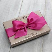 A closed eight piece assortment box. The lid of the box is a satin gold. The box is tied with a large fuchsia ribbon.