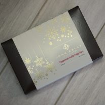 A closed eight piece peppermint chocolate box. The box is a dark brown and has a white sleeve on it. The sleeve is adorned with gold, elegant snowflakes.