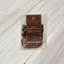A clear container sits with a dark brown label on it. Inside the container is square pieces of the dark chocolate bark. Orange and cranberry bits can be seen outside of the bark.