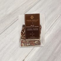 A clear container sits with a dark brown label on it. Inside the container is square pieces of the milk chocolate bark. Almonds and caramel can be seen outside of the bark.