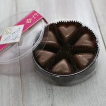 An open plastic, clear, round container. The container holds six milk chocolate hearts. The lid of the container is leaned against the container. The lid has a fuchsia label on it that has a John Kelly Chocolates logo on it.