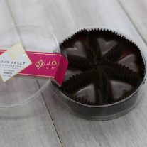 Six dark chocolate hearts sit in a clear, plastic, round container. The lid of the container is leaning against the container. A fuchsia label with a John Kelly Chocolates logo can be seen on the lid.