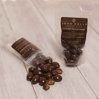 Two bags of milk chocolate almonds sit. One is standing up with the brown label facing outwards. The other is laying down with the brown label facing up, most of the milk chocolate almonds can be seen falling out of the bag.