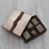 An open six piece assortment box. Various chocolates from the assortment can be seen. The lid of the box is partially on the box, The lid is a light brown and has a dark brown ribbon horizontally across it.