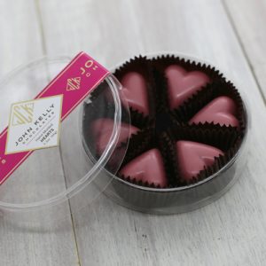 Six pink ruby chocolate hearts sit in an open plastic, clear, round container. The lid of the container is leaned against the container. The lid features a pink label on it with a John Kelly Chocolates logo.