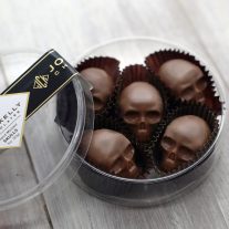 An open container with five pieces of milk chocolate skulls in it. The container is a clear, round, and plastic. It has a black John Kelly Chocolates logo on it.