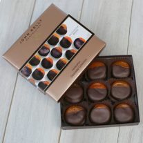 An open box of the chocolate dipped apricots sits with the box covering one of the nine apricots. The apricots are all sitting neatly in paper, brown cups. The apricots are mostly covered in chocolate leaving the top one fourth uncovered. The lid has a white sleeve on it with a picture of the apricots on it.