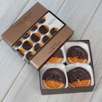 An orange slice box sits open with the lid partially on the box. The orange slices are mostly covered in chocolate with a fourth of the orange uncoated. The lid of the box is a light brown and has a white sleeve on it with pictures of the orange slices on it.
