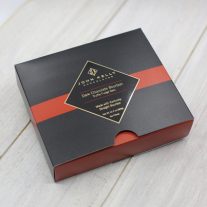 A black, square box with a red stripe going horizontally through the middle. The stripe has a black John Kelly Chocolates logo on it. There is a semi-circle at the bottom of the box that shows part of the red box that slides out of the black box.