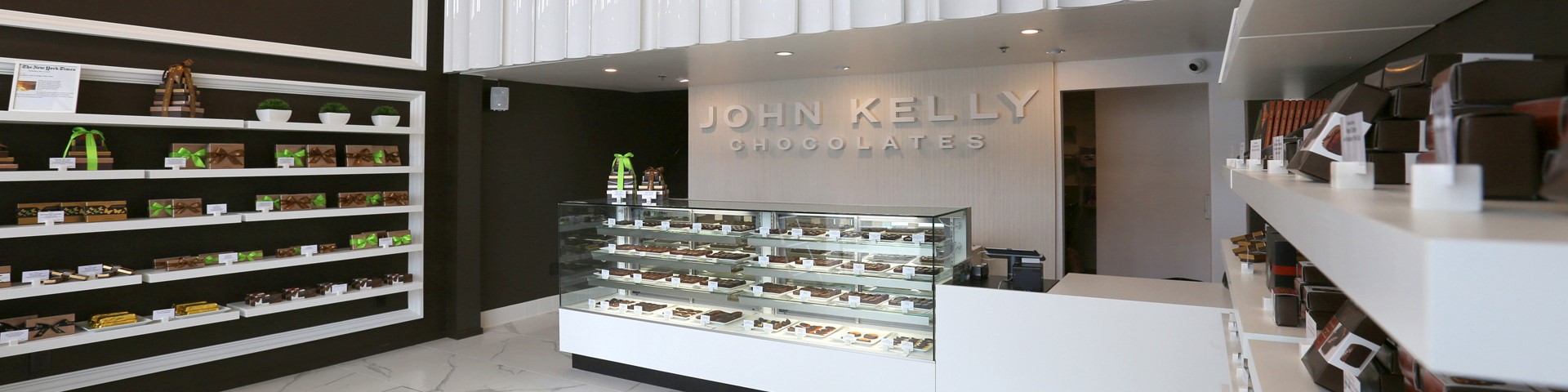 A picture of the front of the John Kelly Chocolates store in Beverly Hills. The colors inside the building are brown and white. A case can be seen with delicious chocolates inside. The walls are lined with towers and assortments of chocolate. A sign in the back reds John Kelly Chocolates.