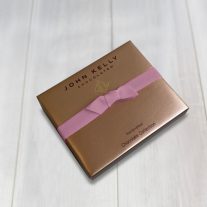 A six piece satin box with a John Kelly Chocolates logo on it. It is closed with an orchid stretch ribbon.
