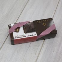A dark brown rectangular box with a red dark chocolate with walnuts sleeve on it. It is closed with an orchid stretchy ribbon.